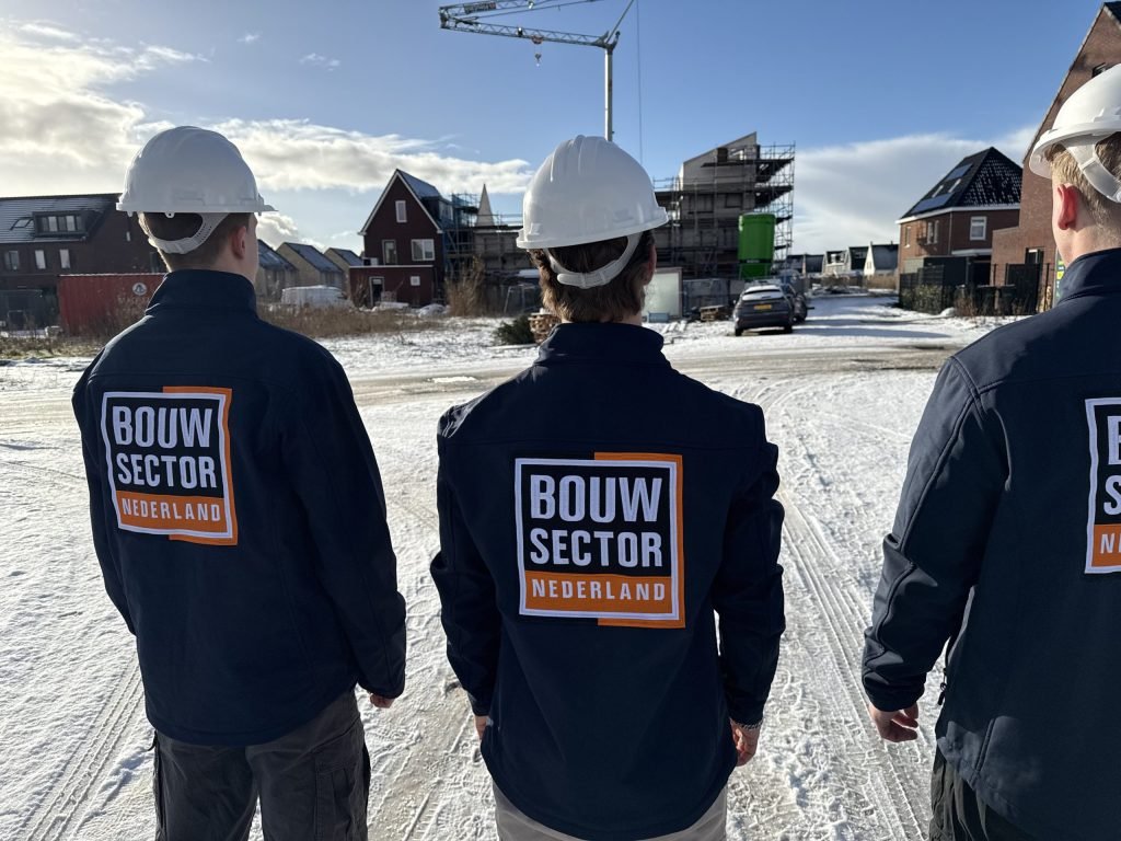 Drie vakmensen van Bouwsector Nederland met witte helm, op de rug gezien bij een besneeuwde nieuwbouwwijk